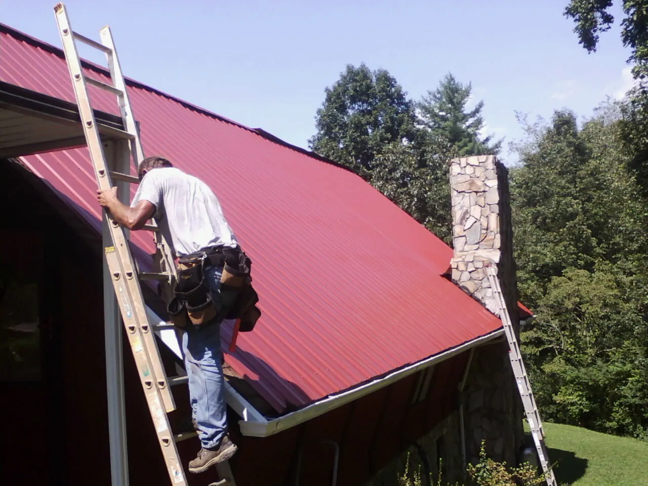 Roofer on a ladder beside a red metal roof and stone chimney — Soffit & Fascia Repair by Roofing Brothers in Capistrano Beach